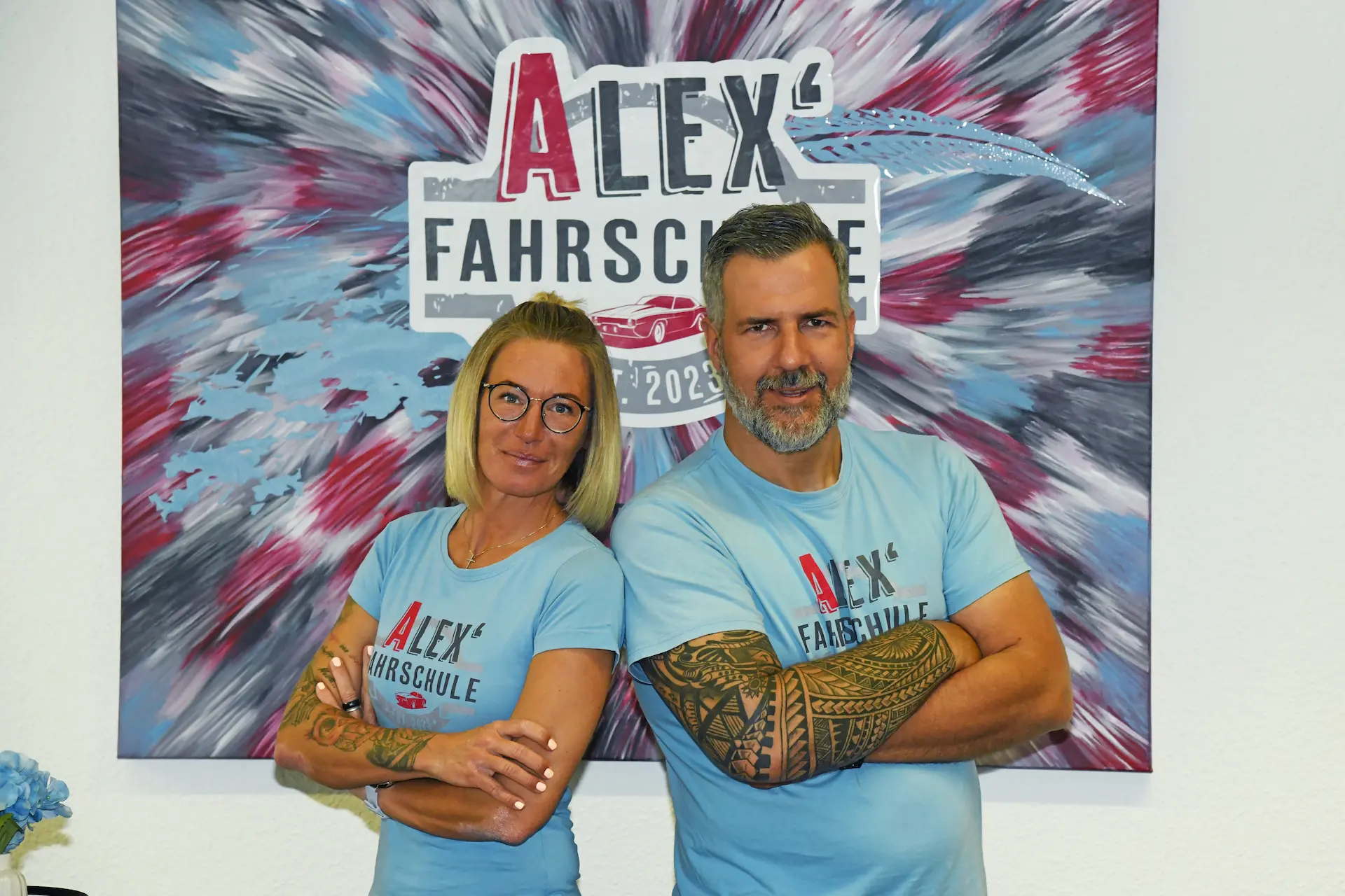 Fahrlehrer Alex und Caro von Alex' Fahrschule vor dem Firmenlogo in Gadebusch